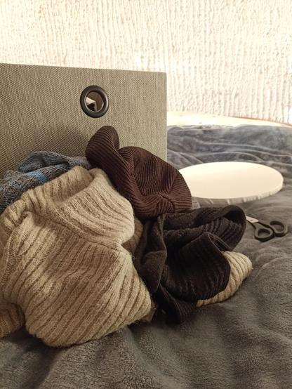 Pile of warm clothes on a grey blanket. Grey box behind the pile. Scissors and empty round canvas are on the background.