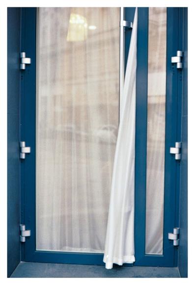 Color photograph of a glass door in a blue door frame. Behind the door is a white curtain, the end of which is tucked between the door and its frame, so that part of the curtain is flapping in the wind outside.