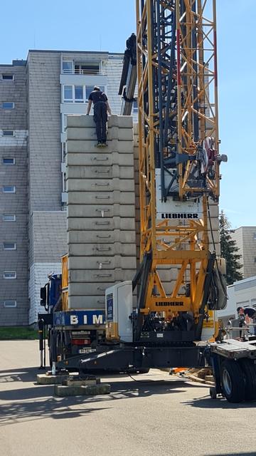 Unterer teil eines baukranes, hoher Stapel von beton-gewichten geladen, die ihn fest an den Boden drücken sollen. Ein Arbeiter ist zum obersten betonblock geklettert, wohl um ihn richtig zu platzieren.
