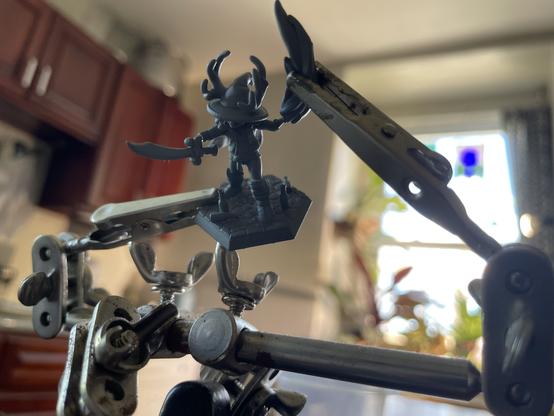 a closeup of a goblin miniature in the process of having his fireball-wielding arm reattached. he's held together with one of those little pinchy magnifying glass armature devices. no idea what it's called.