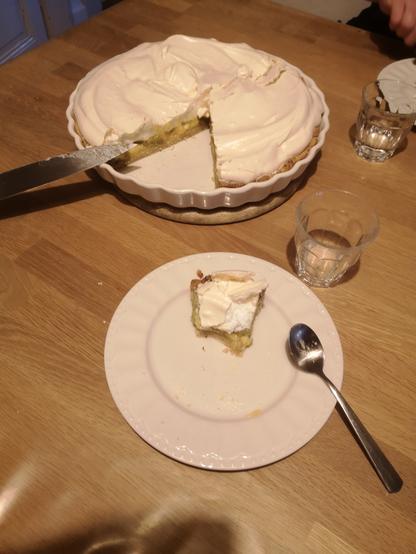 Photo d'un plat à tarte rond avec dedans une tarte à la rhubarbe meringuée. 1/6 de la tarte est coupé et une part entamée est présente sur une petite assiette à coté.
Un grand couteau est posé sur le plat à tarte.