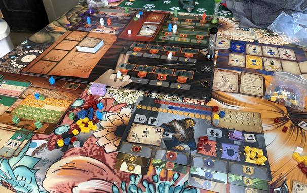 The Pendulum board game is setup on a table with a floral pattern tablecloth. The player’s board is closest to the photographer with the Automa board next to it, both surrounded by player tokens. The main board is further back and has an array of worker meeples on action spaces. Cards are missing as the game is being cleaned up.