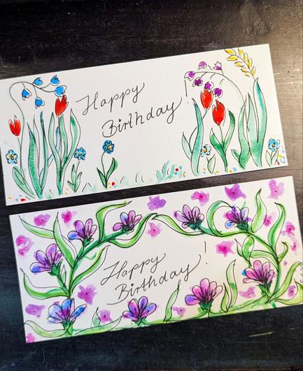 Schwarze Tischplatte, darauf 2 längliche Geburtstagskarten übereinander. Beide mit floralen Motiven, die mit schwarzem Stift nachgezeichnet sind. Bei beiden steht in der Kartenmitte "Happy Birthday!" - wobei der Punkt über dem "i" als winziges Herz gemalt ist.
