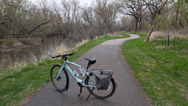 Sage green Vado SL ebike parked on the Bear Creek trail with the creek on the left and green grass on either side of the trail with trees surrounding now with green leaves emerging under cloudy skies.