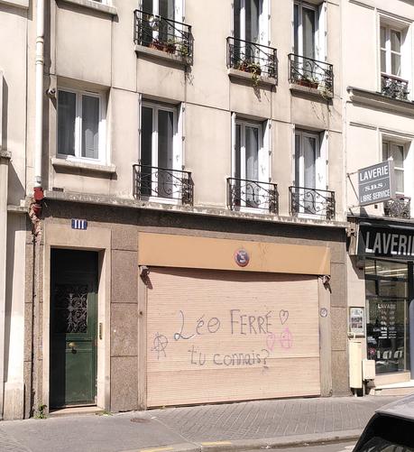 Immeuble parisien avec une porte d'entrée et un rideau de fer sur un garage, portant l'inscription "Léo Ferré tu connais ?" avec des coeurs et des A entourés (anarchiste)
