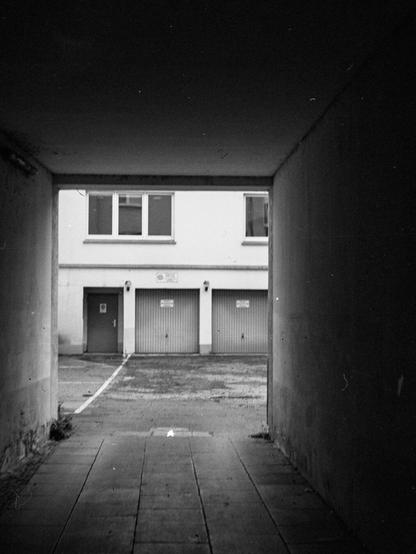 Black and white photo of a backyard with some garages, seen through a dimly lit tunnel. There is some noticable motion blur from camera movement.