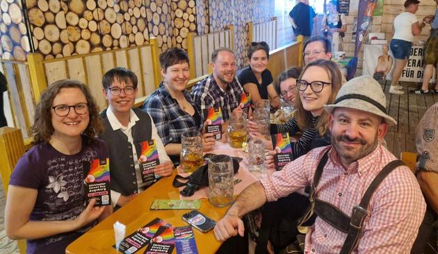 Mitglieder des CSD-Vereins an einem Biertisch im Zelt. Ganz rechts mein grüner Kollege Peter Rauscher in Tracht. Ich ganz links. Viele halten Flyer für den Augsburger CSD 2025 am 14. Juni in der Hand.