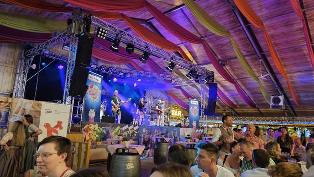 Ein Blick übers Schaller-Festzelt. Unten im Bild sitzen Leute an Biertischen. In der Mitte sieht man eine blau beleuchtete Bühne. Die Decke ist in rosa-lila beleuchtet. Auf der Bühne spielt eine Band.