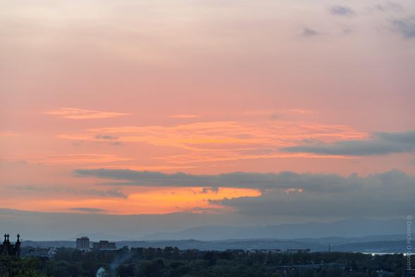 A tranquil sunset scene over Edinburgh, Scotland, with soft pastel hues of pink, orange, and lavender filling the sky above distant hills and silhouetted city buildings; faint smoke rises from the greenery below.