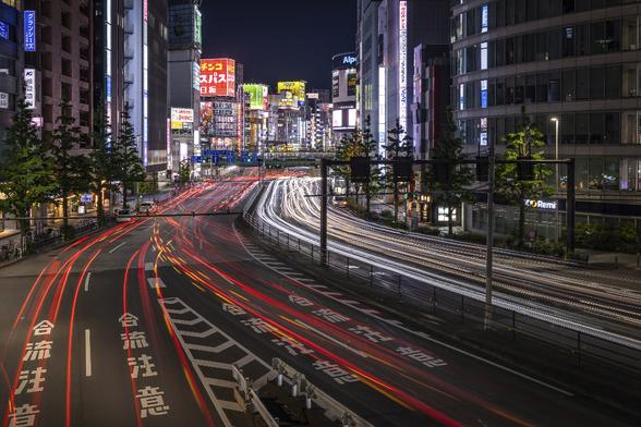 Long exposure shot on a buzzy main road of Shinjuku in Tokyo from a bridge over the road, at night. Only the light trails of the cars are visible on the road in the foreground due to the long exposure. Skyscrappers and buildings covered in colourful neons are recognisable in the background.