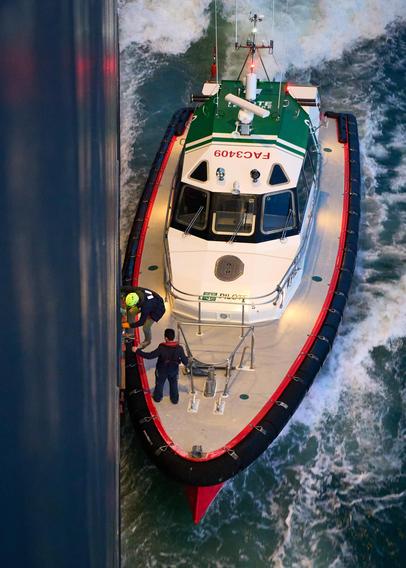 The image shows a small pilot boat with a green and white cabin navigating along the side of a larger ship. Two individuals are on the deck, one in a helmet gesturing towards the larger vessel. The sea is choppy, creating splashes of white water around the boat.