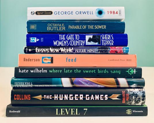 Pictured is a stack of books including George Orwell's 1984, Octavia Butler's Parable of the Sower, Sheri Tepper's The Gate to Women's Country, Aldous Huxley's Brave New World, M.T. Anderson's Feed, Kate Wilhelm's Where Late the Sweet Birds Sang, Frederik Pohl and C.M. Kornbluth's The Space Merchants, Suzanne Collins's The Hunger Games, and Mordecai Roshwald's Level 7.