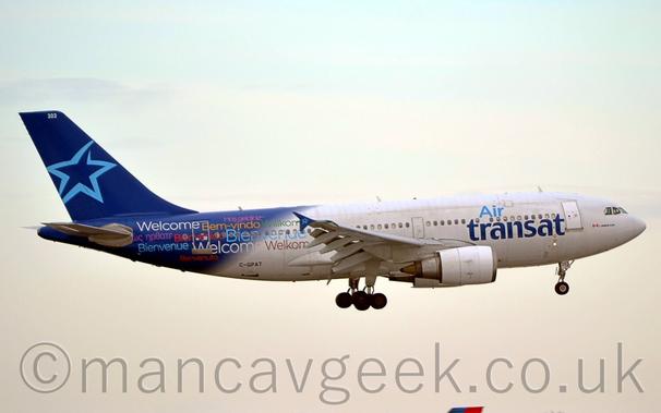 Side view of a white and blue , twin engined jet airliner flying from left to right at a very low altitude, with undercarriage lowered and flaps extended from the rear of the wings, suggesting it is about to land.
The plane is mostly white, with blue "Air Transat" titles on the forward fuselage.
The rear fuselage is dark blue, with the pale blue outline of a star on the tail.
Greetings in various languages and colours are scattered across the rear fuselage - "Welcome", Welkom", Bienvenue", and many more similar sentiments.
The registration "C-GPAT" is on the lower rear fuselage in black.
Pale grey sky fills the rest of the frame.