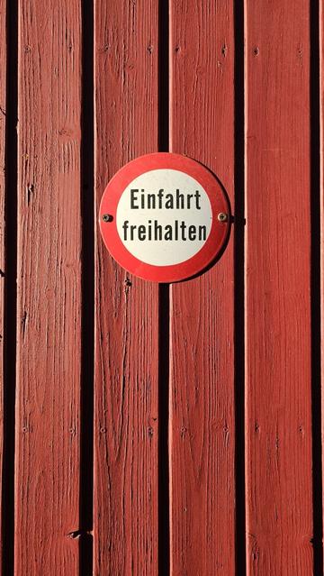 The image shows an oval sign mounted on a red wooden surface. The sign has a white background with a red border. Black text is present on the sign, reading "Einfahrt freihalten".