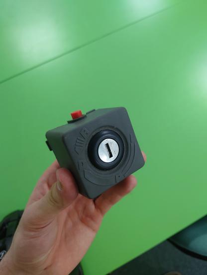 The original battery connector in my hand, showing a small grey box, with a car style key ignition switch, and a red button on the top.