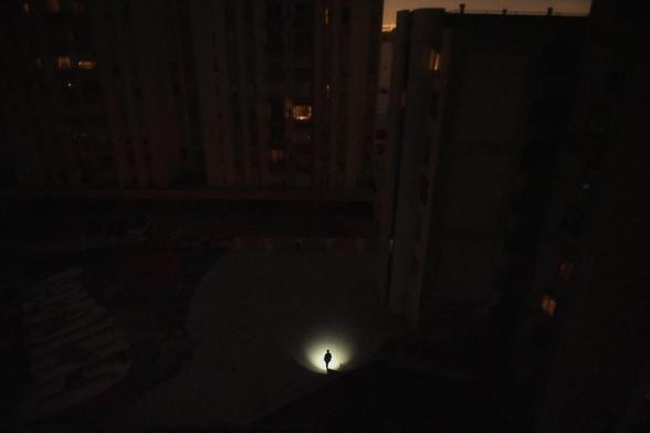 A solitary figure stands illuminated by a flashlight in the middle of a dark urban courtyard surrounded by tall buildings at night.