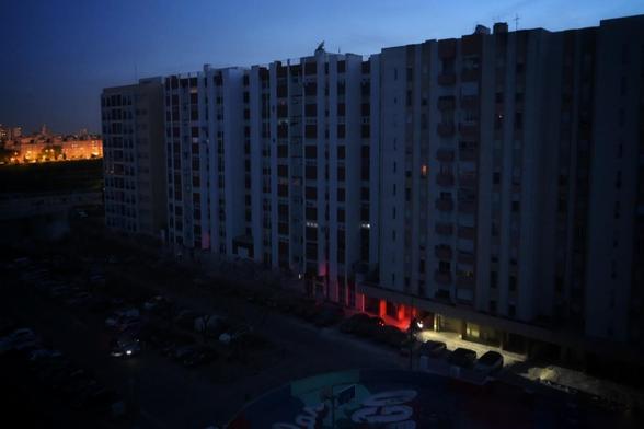 A large, multi-story residential building is illuminated by dim, colorful lights with a cityscape in the early evening sky.
