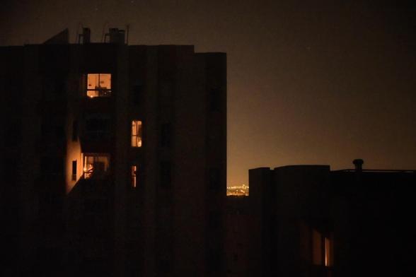 A dimly lit night scene shows a building with a few illuminated windows, set against a faintly glowing city skyline.