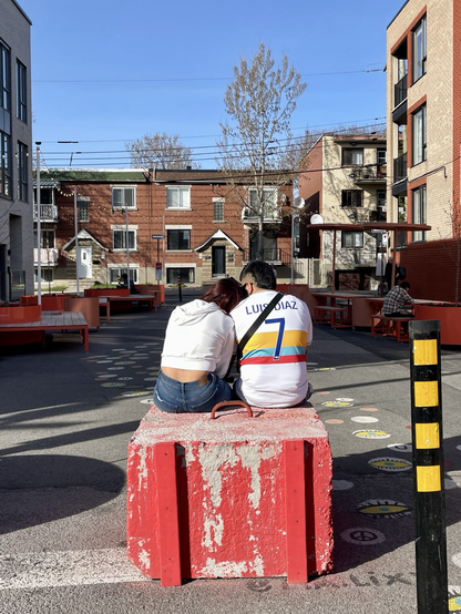 A couple sits affectionately on a cement block at the entrance of a pedestrian section of a city street.