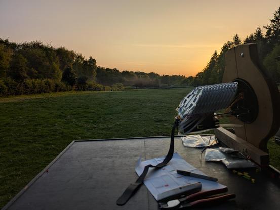 A wooden table in a large field surrounded by forest. A golden sunset is in the sky. Some work equipment is on the table.