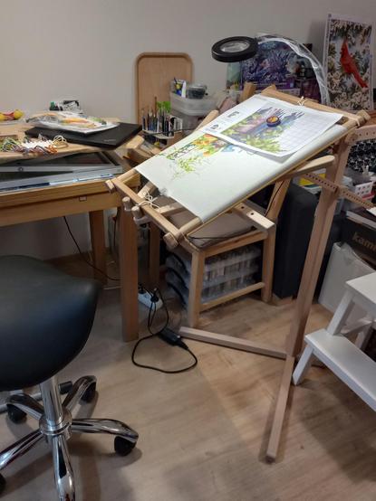 Cross stitch piece in progress stretched on a wooden frame that is resting on a wooden floor stand. A paper pattern chart is attached to the canvas with a magnet. The work is illuminated by a ring lamp clipped to the stand.