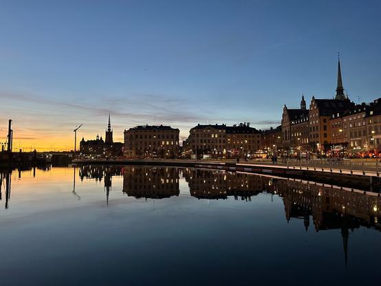 buildings on the southwestern shore of Gamla Stan, the old town of Stockholm, reflecting in the water of the lake at dusk. The orange glow of the recently set sun is visible on the horizon to the left, over the water