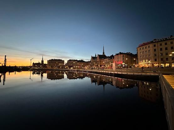 a wide-angle view of buildings on the southwestern shore of Gamla Stan, the old town of Stockholm, reflecting in the water of the lake at dusk. The orange glow of the recently set sun is visible on the horizon to the left, over the water