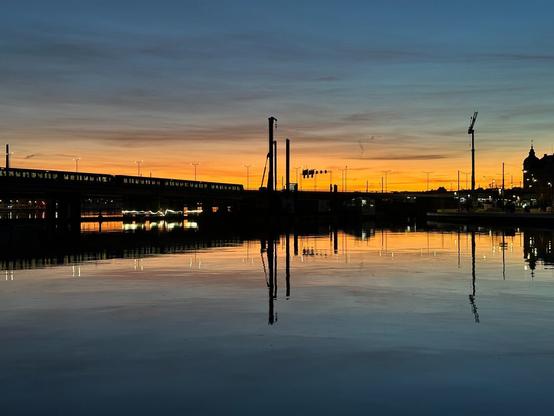 a view over water towards the silhouette of a bridge backlit by a golden sunset. The lights in the windows of a passing metro train are visible over the bridge on the left. Below them, in a gap under the bridge is the white squiggle of industrial LED ribbon lighting, as used on construction sites