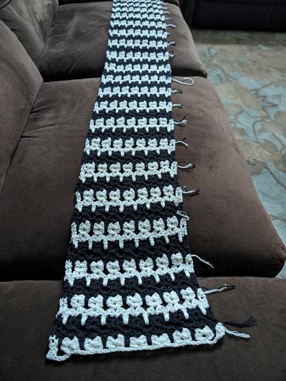 black-and-white tessellated cat scarf on brown couch - one side is fully finished, the other side still has yarn ends sticking out between every pair of cat rows