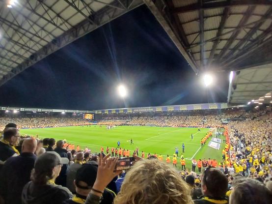 A full elland road at night. There are stewards round the edge of the pitch to prevent an invasion. The crowd is all in yellow. The pitch is empty and getting ready for the Leeds players to come out for promotion celebrations