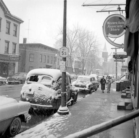 A view down a street in Fort Plain, New York. Cars are parked along both sides of the street. Visible business signs include: "Dobber's Toggery", "Western Auto Associate Store", and "Ron-D-Voo Inn". On a lamp post is a road sign that says "N.Y. 5s".