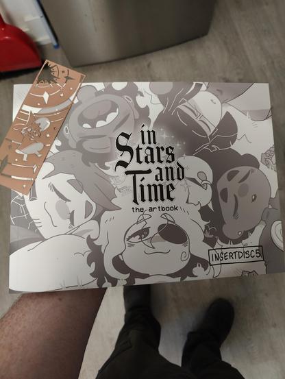 A photo of a large art book for In Stars and Time. It has black and white illustrations of the cast in the front. Sitting on it is a copper filigree boomark