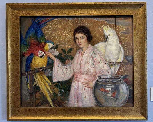 photo of the painting in gold frame on display at museum: oil painting in landscape orientation, portrait of a woman with fair skin and short bobbed dark hair in a pink robe posing with two macaws on perches to the left (appear to be a Blue and Gold Macaw and a Green-Winged Macaw) and a white cockatoo on another perch to the right (appears to be either a Yellow-Crested or Sulfur-Crested Cockatoo), with a fishbowl with two orange goldfish also next to her in lower right corner, against a sparkly golden background.