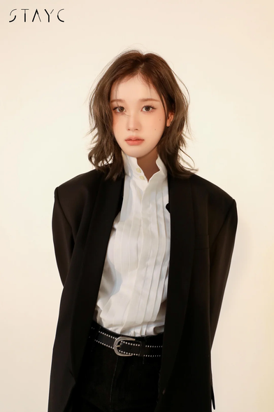 The image features  Kpop idol J standing against a plain, light-colored background. She is wearing a black blazer over a white shirt with vertical pleats, which adds texture and detail to the outfit. The shirt is buttoned up, and the blazer is open, creating a relaxed yet polished look. J is also wearing a black belt with a silver buckle, which has a studded design, adding a touch of edginess to the ensemble. The belt cinches the waist, complementing the high-waisted black pants. The hair is styled in loose waves, falling just past the shoulders. The image includes the text "STAYC" in the upper left corner, indicating the group to which J belongs.

Provided by @altbot, edited by @jemmesedi