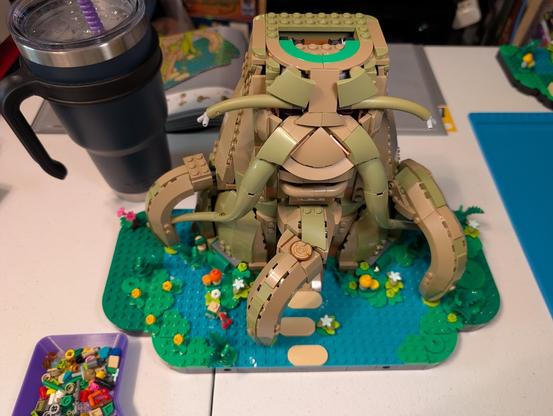 Photo of a partially assembled Lego set (77092 Great Deku Tree 2-in-1).

The instruction book is open at the top but the contents cannot be seen.

The visible portion of the partially assembled set has a mostly complete trunk and a wide base that is green and teal-ish and has numerous small flowers and plants around. The trunk has parts resembling a face with eyebrow and a mustache-like vines, eyes, a nose, and mouth. Three thick roots extend from the trunk and out onto the base.

There is a large metal tumbler full of water with a purple straw off to the left.

There is a small purple sorting tray near the bottom left.