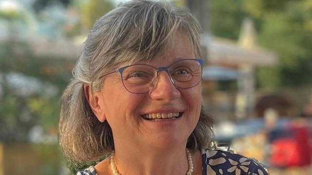 Portrait von Andrea Petzold. Eine Frau mittleren Alters mit grau melierten, schulterlangen Haaren. Sie trägt eine Brille mit blauen Bügeln und ein dunkelblaues, gemustertes Oberteil. Sie lächelt breit und wirkt fröhlich. Der Hintergrund ist unscharf und zeigt einen Außenbereich mit etwas Grün.