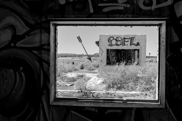 A view out of an abandoned building's window. The photo is in black and white. The inside walls are covered in graffiti. Outside there is a small box like structure with a large window, but no glass. Across the top is graffiti that reads, BSFL. On the left, further out is a large arrow positioned such that it appears stuck into the ground. It is probably twelve feet long and stuck into the ground at a thirty degree angle.