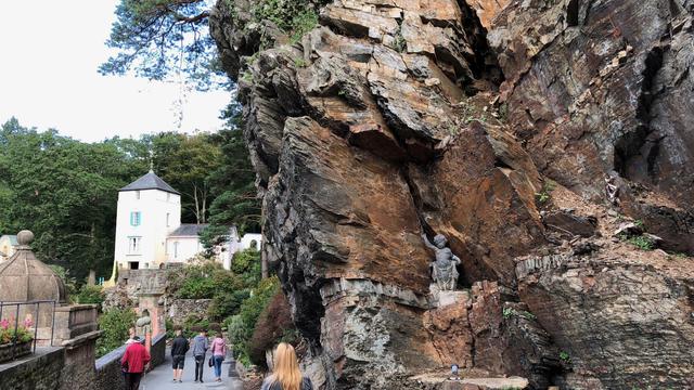 This picture captures a scenic view from Portmeirion, a tourist village in Gwynedd, Wales. The image shows a narrow pathway lined with stone walls and lush greenery on both sides. On the right, there is a striking rugged rock formation with a small statue of a child-like figure embedded in a niche within the rocks. The left side of the image reveals charming, pastel-colored buildings, including a distinctive white tower with teal window shutters and a conical roof, nestled among dense green trees. Several people are walking along the path, adding life to this picturesque and tranquil setting.