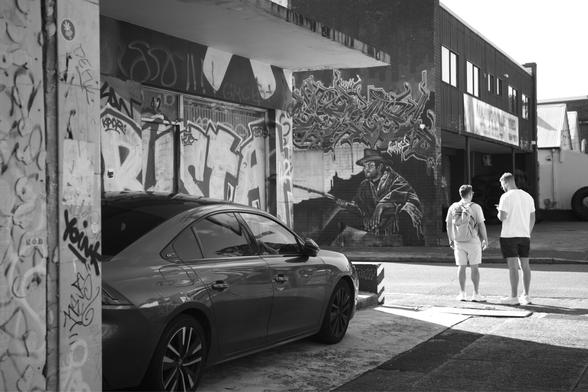 Black and white shot of two guys figuring out how to get where there going. Tons of street art dots the light industrial buildings near them. They stand in sunlight.