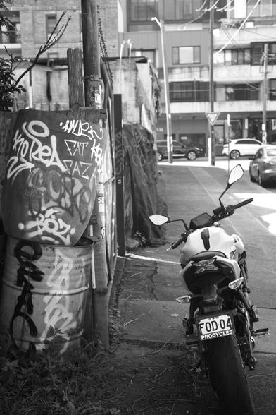 Black and white shot of a bike parked by some street art the back of an abandoned lot. In the distance some new apartment complexes are visible.