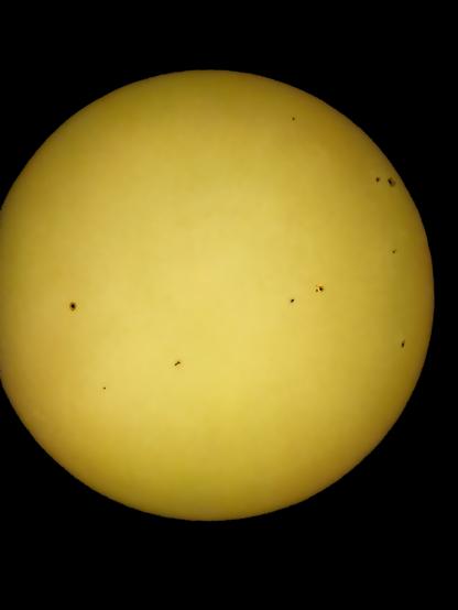 „Fotografie der Sonne vom 27. April 2025, aufgenommen mit Sonnenfilter und Telekonverter, zeigt mehrere dunkle Sonnenflecken auf der Sonnenscheibe.

Photograph of the Sun taken on April 27, 2025, using a solar filter and teleconverter, showing several dark sunspots across the bright yellow disk.