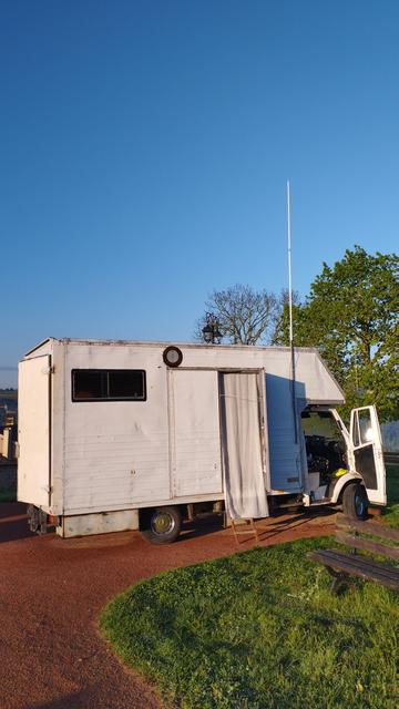 Le tawamusi à l'arrêt sur un parking en graviers rouges. Une antenne de 2m dépassant du toit sur le côté droit. En arrière plan des arbres et un ciel bleu