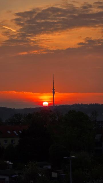 Der Fernsehturm Dresden steht im Zentrum der Aufnahme. Die Sonne berührt gerade den Horizont, der Himmel ist teils wolkig und in warme Farbtöne getaucht. Im Vordergrund erkennt man Bäume und Hausdächer im Morgendunkel.

The Dresden TV tower stands in the center, with the sun just above the horizon. The sky is partly cloudy and painted in soft morning colors. Trees and rooftops are visible in the shadowy foreground.