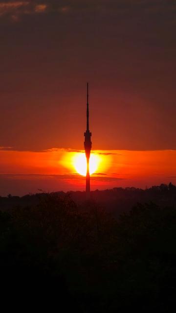 Die Sonne geht genau hinter dem Fernsehturm Dresden auf und bildet eine symmetrische Silhouette. Die Umgebung liegt im Dunkeln, während das intensive Licht der Sonne in Orange- und Rottönen durch die Wolken bricht.

The sun rises directly behind the Dresden TV tower, creating a symmetrical silhouette. The sky glows with warm orange and red hues, while the foreground remains in darkness.