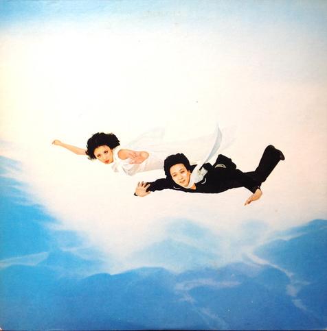 The cover art is a photo of two people (Mika and Kazuhiko Katō, I think) who appear to be flying in the air, a landscape seen below them and white clouds behind them, arms and legs spread out, facing the ground. One is wearing black pants and a black shirt, with a white scarf flapping in the wind behind them. The other is wearing all white. They both have longish hair flying behind them, and are looking at the camera.