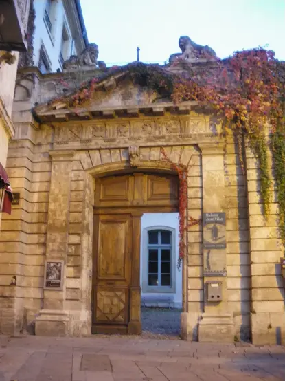 The image features an ornate door set within a stone archway, which has decorative carvings and details on the facade. There is greenery, likely vines, climbing along the top of the arch. The entrance appears to lead to a courtyard or garden area, as suggested by the window visible in the background. The overall style suggests a historic or architectural significance, typical of old European buildings.

Image Credits: Wikimedia / Véronique PAGNIER / Public domain