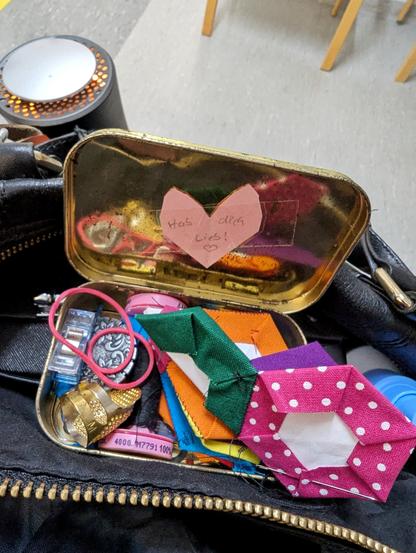 Grauer Wartezimmerfußboden. Eins sieht eine offene schwarze Tasche, daran hängt ein gelb leuchtender Luftfilter. In der Öffnung steht ein offenes Blechdöschen mit allerlei Nähezubehör und bunten Stoffhexagonen, die zusammengenäht werden.