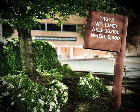 A tree, hedges, a bush, and a brown metallic sign in the foreground frame a coral-colored building and its parking lot at night in the blurry distance. The sign reads, "TRUCK WT. LIMIT | AXLE 10,000 | WHEEL 5,000."