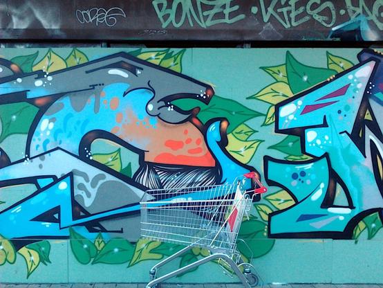 Ein leerer Einkaufswagen steht vor großflächigen Graffiti an einer hässlichen Wand in der Ruine des Ihmezentrums in Hannover-Linden.