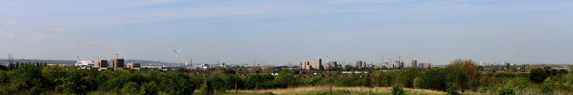 Wide image showing a wooded foreground with a horizon formed of urban development, prominent amongst which are new mid rise apartment buildings and a number of wind turbines.